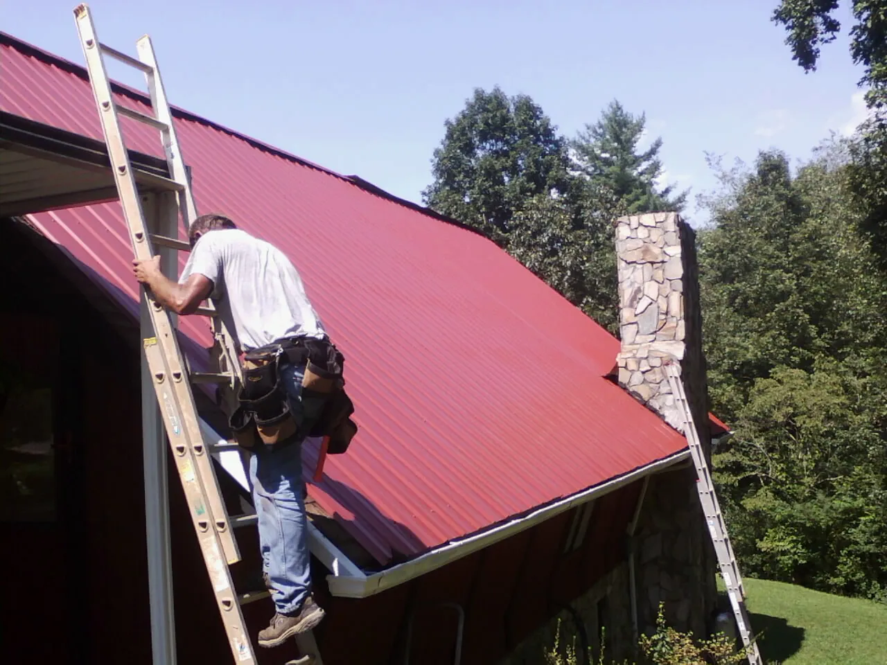 Roofer on a ladder beside a red metal roof and stone chimney — Soffit & Fascia Repair by Roofing Brothers in Cottekill